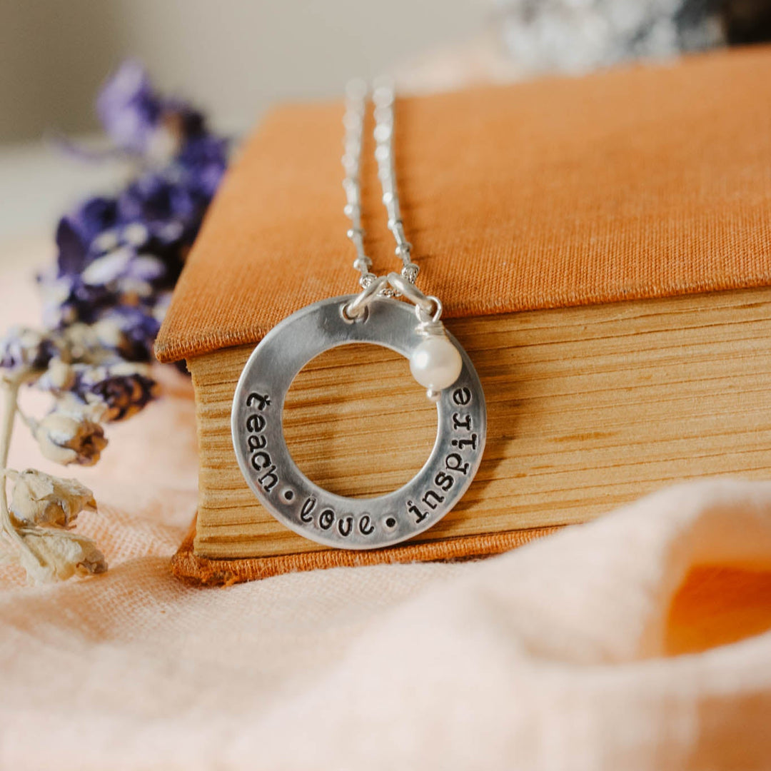 Silver necklace with a circular pendant on a wooden surface with flowers and fabric.