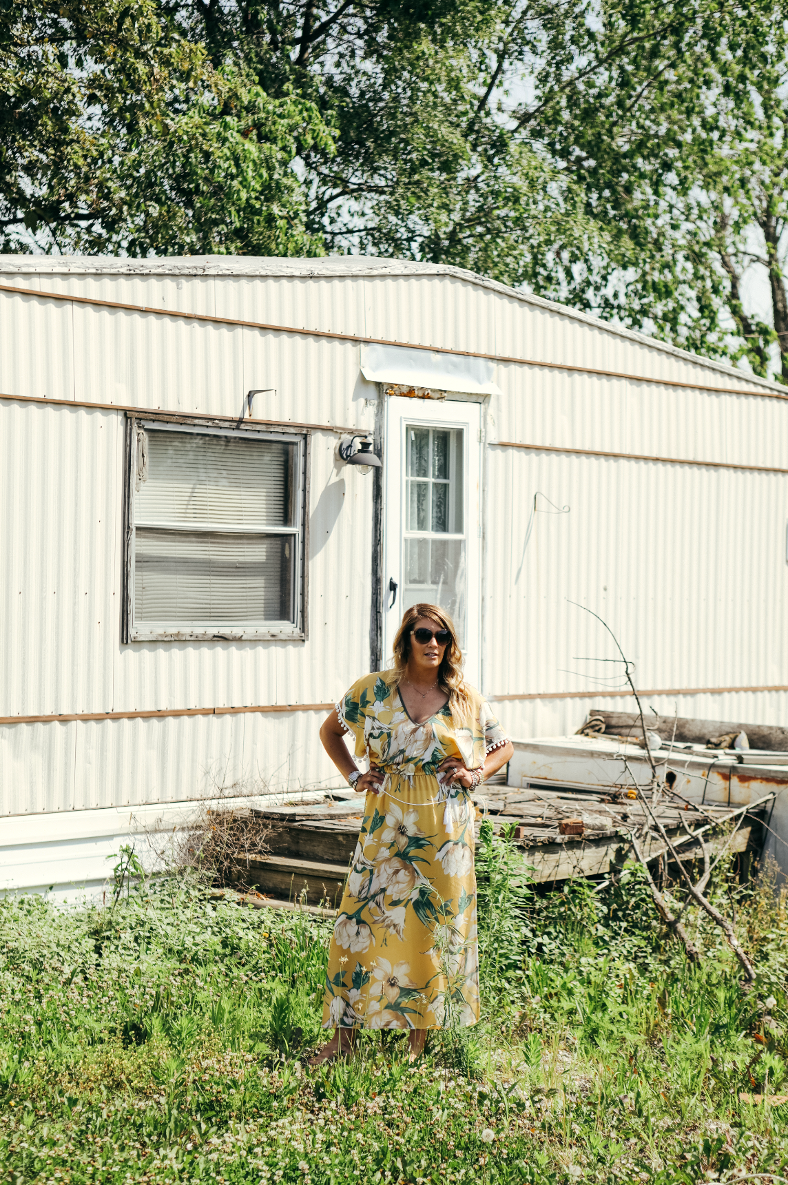Woman in a yellow floral dress standing in front of a mobile home with overgrown grass and weeds.